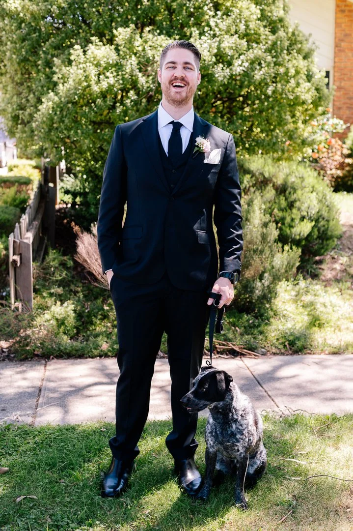 A man in a dark suit with a white shirt and black tie stands outside on a sidewalk, smiling, holding a leash attached to a brindle dog, in front of a lush green bush and a residential area.