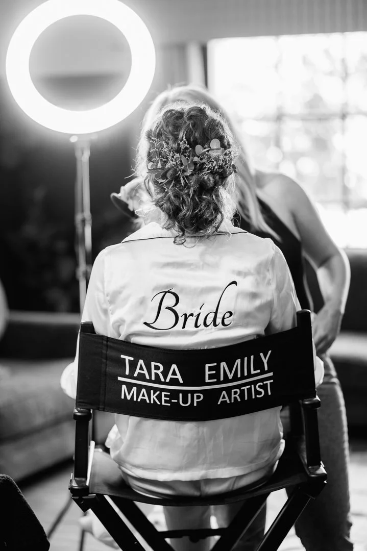 A bride with hair decorated with flowers sitting in a makeup artist's chair, with a bright ring light illuminating her from behind.