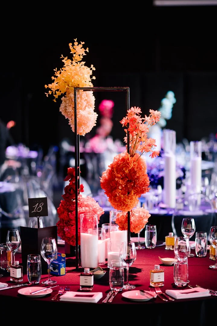 Elegant banquet table with tall floral centerpieces in shades of pink and orange, surrounded by candles, glassware, and place settings, in a dimly lit event hall.