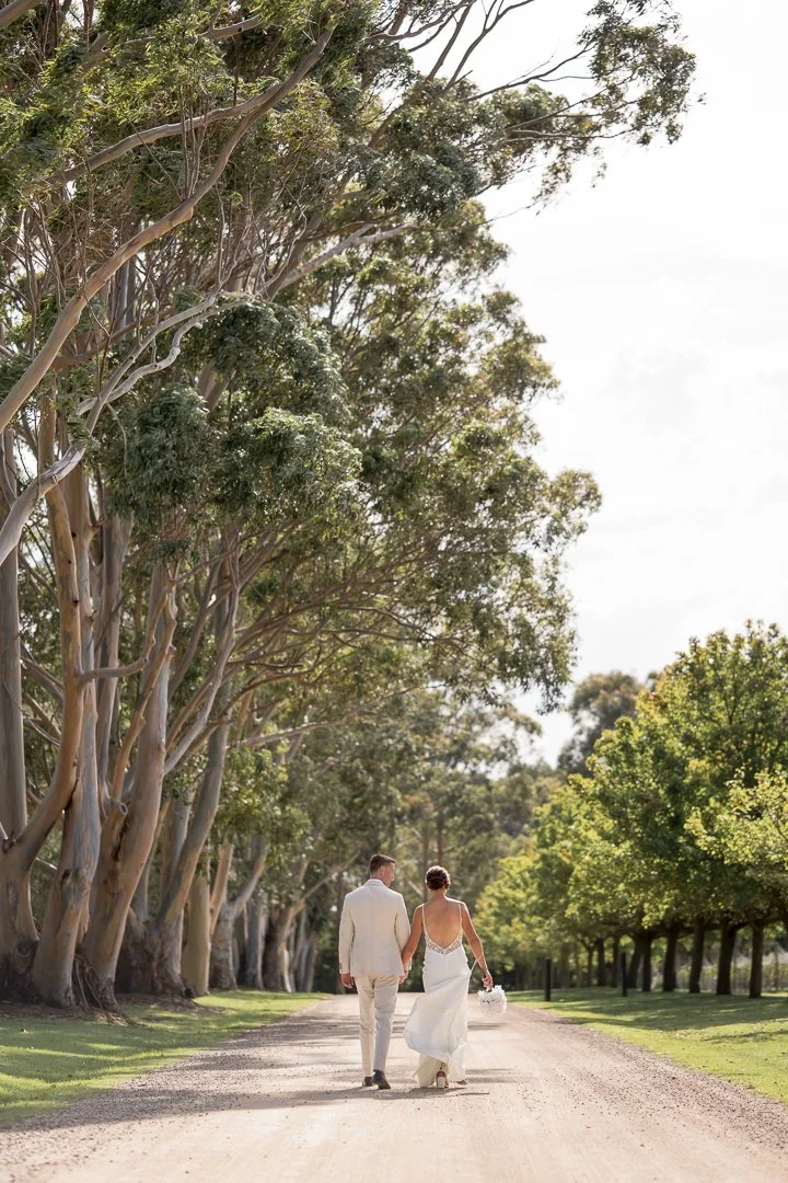 A couple dressed in wedding attire walking hand in hand on a dirt road surrounded by tall trees and lush green foliage.