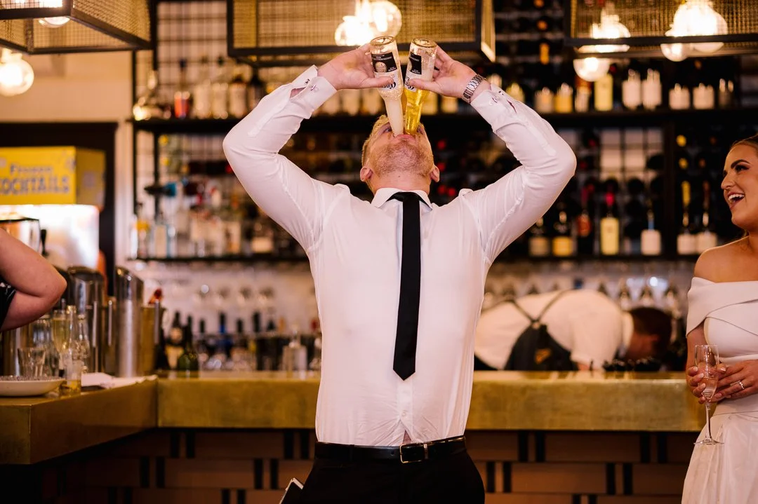 A man wearing a white shirt and black tie drinks two bottles of beer at the same time in a bar. A woman in a white dress holding a glass of champagne smiles nearby.