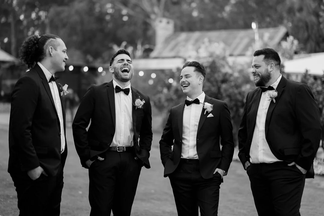 Five men in tuxedos standing outdoors, laughing and talking casually at a formal event or wedding.