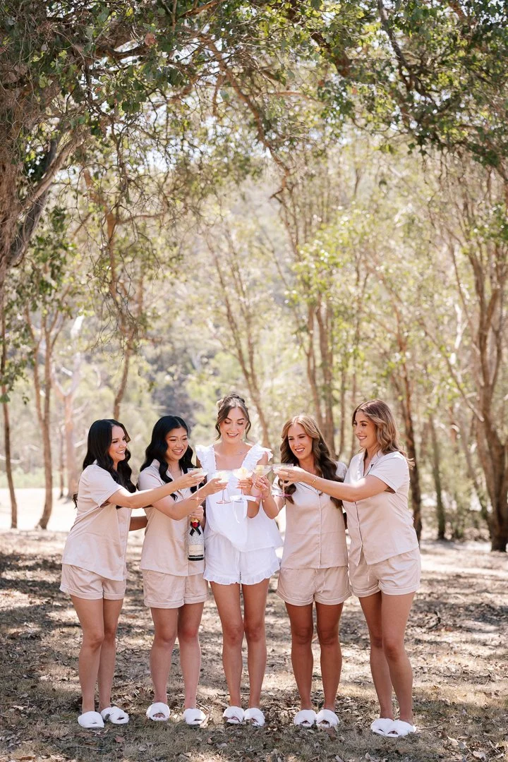 A group of five women dressed in light-colored pajamas and slippers, standing outdoors in a wooded area, celebrating with drinks raised in a toast.