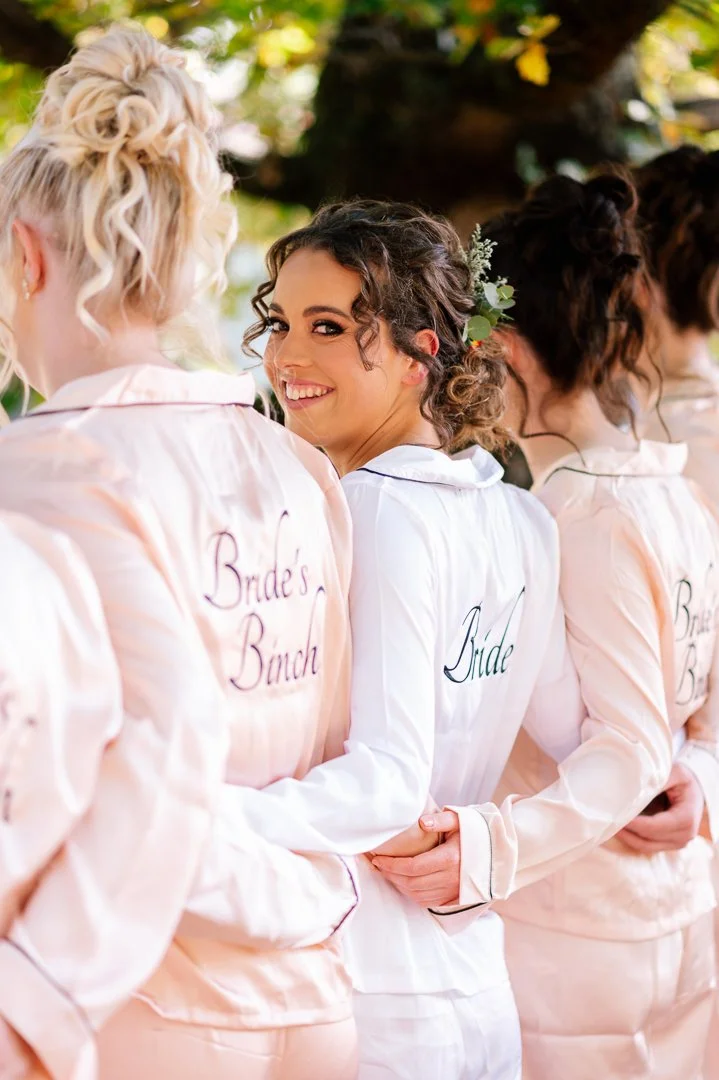 A group of women in satin pajamas with 'Bride' and 'Bridesmaid' embroidered on the back, standing together outdoors. One woman, the bride, is smiling at the camera with dark, curly hair and a green hair accessory.