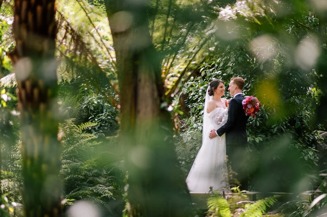 Bride and groom standing close together, smiling, surrounded by lush greenery in an outdoor setting.