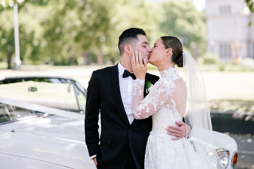 A bride and groom sharing a kiss outdoors on their wedding day, with a vintage white car in the background.