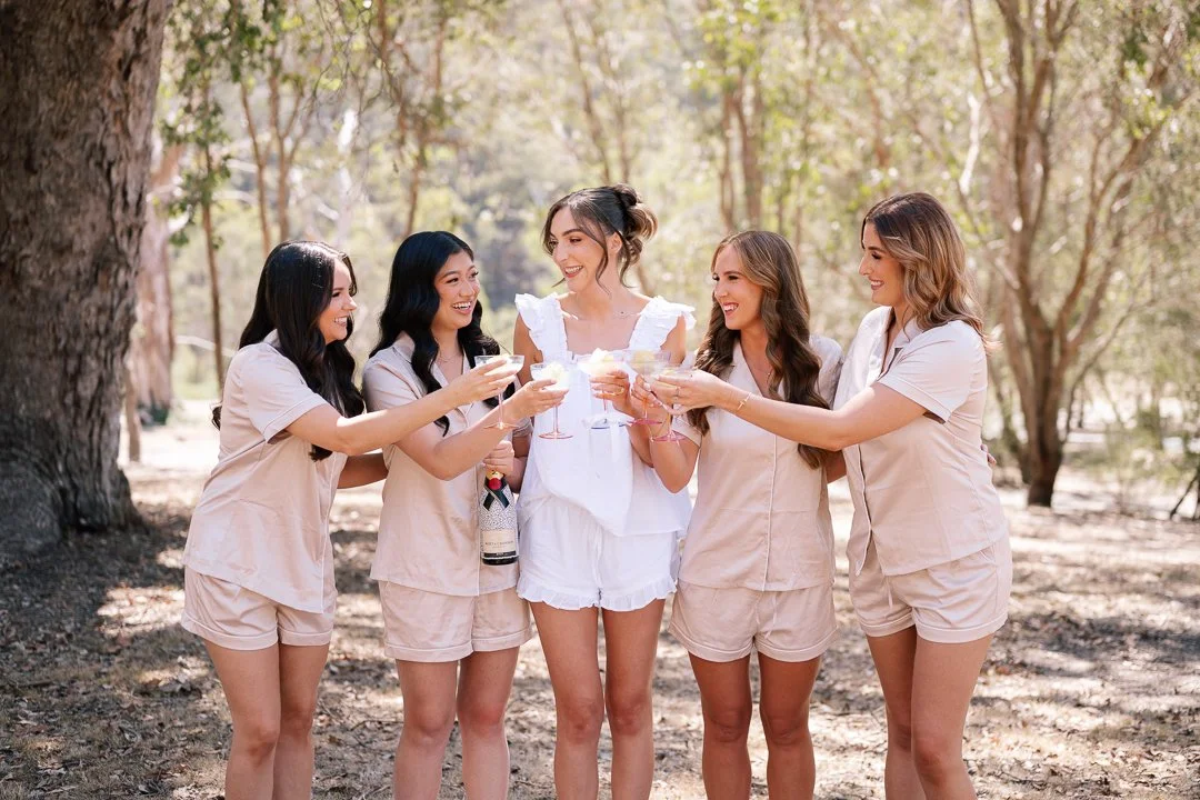 Five women in casual beige outfits celebrating outdoors with drinks, smiling and toasting in a shady wooded area.