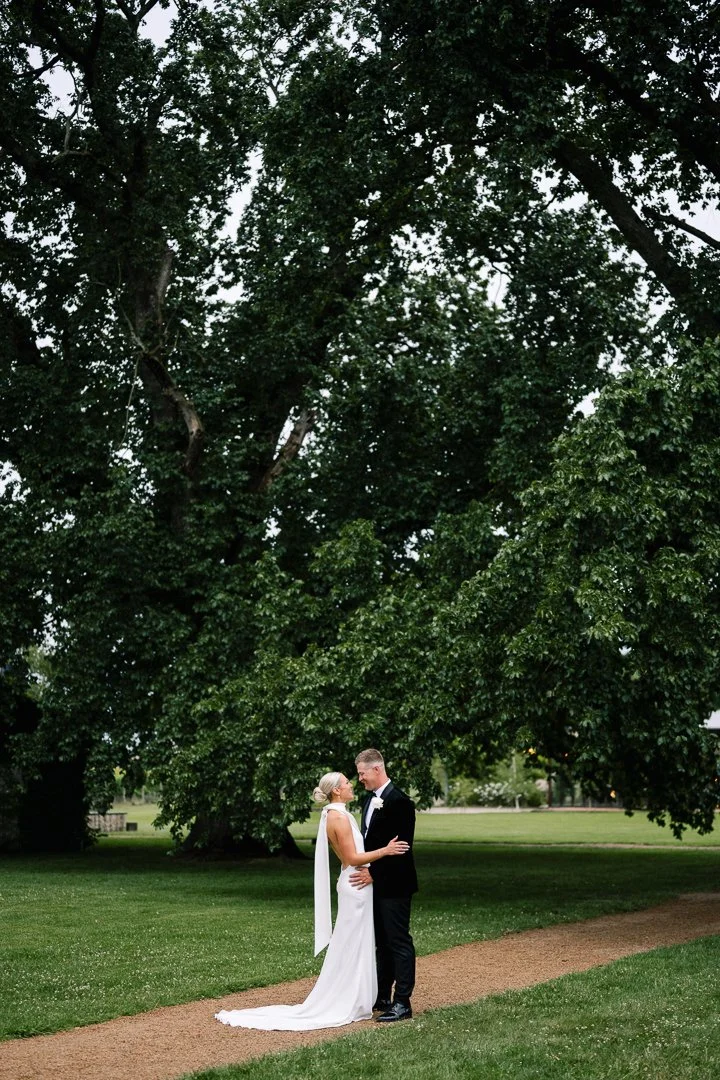 A bride and groom stand facing each other on a park path, embracing and smiling, with a large green tree in the background.