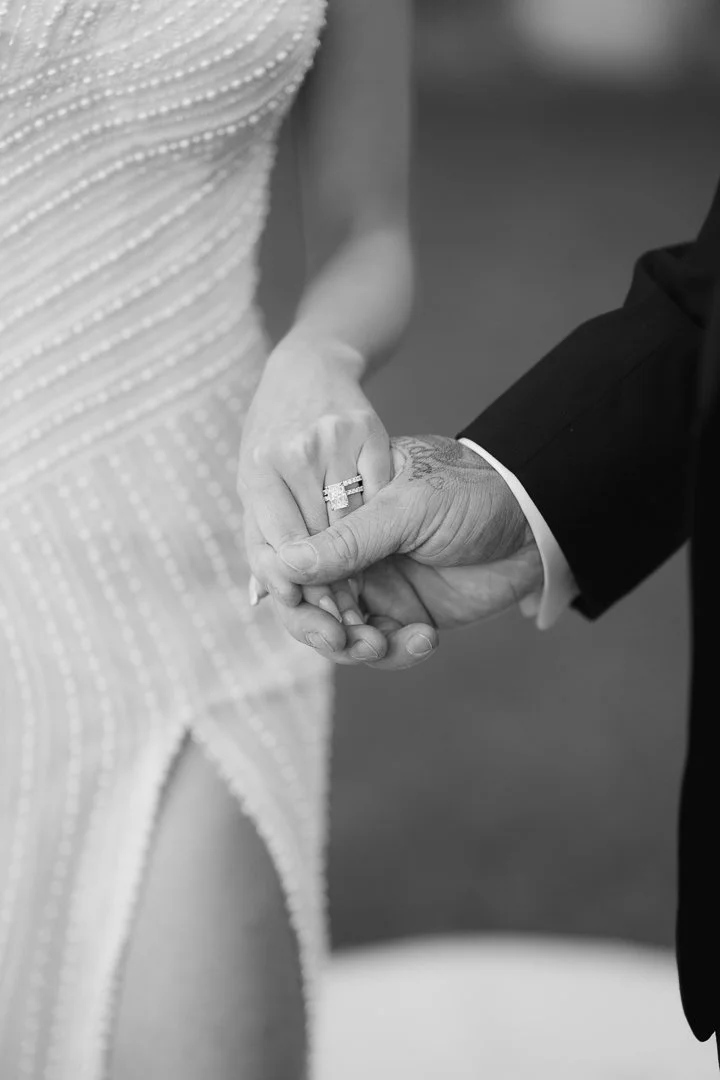 Close-up of a bride and groom holding hands during a wedding, focusing on the bride's wedding ring and the groom's hand, with the bride wearing a dress and the groom in a suit.