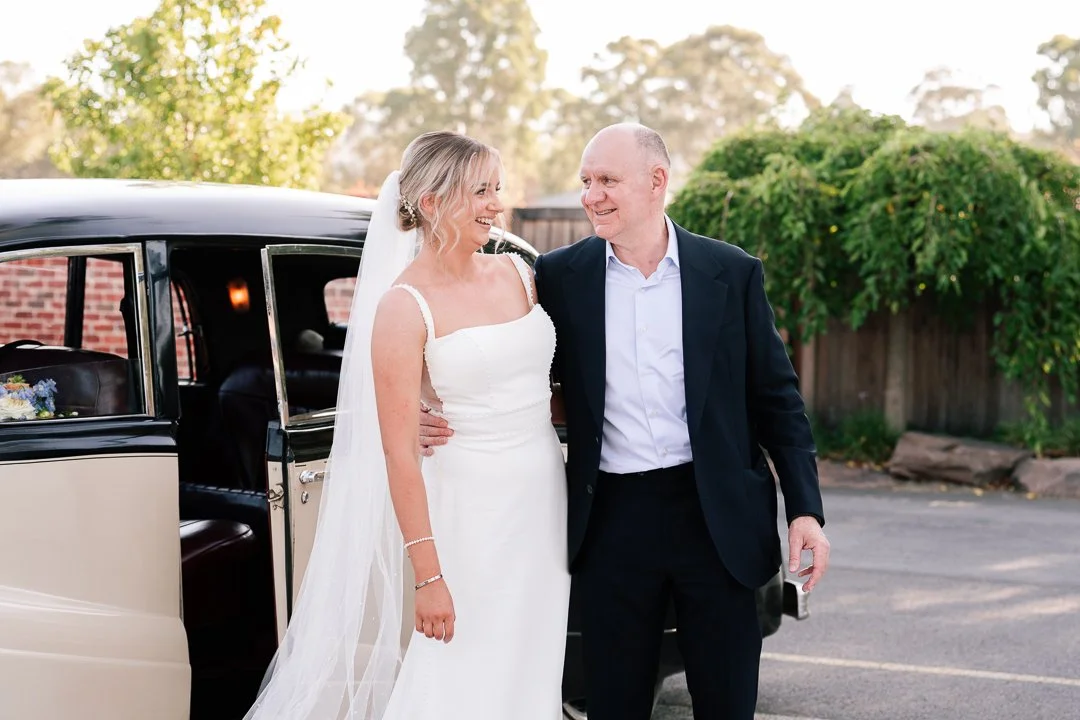 A bride in a white wedding dress with a veil smiling and standing next to a man in a black suit, in front of a vintage car outdoors during daylight.