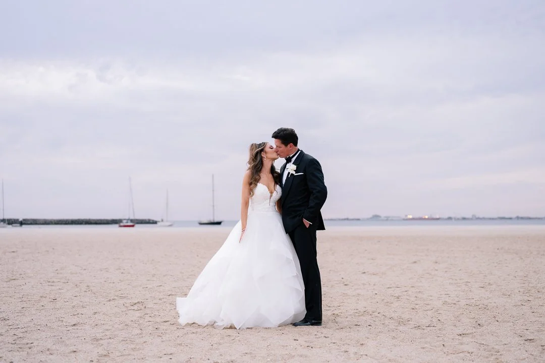 A bride and groom kiss on a sandy beach with calm water and sailboats in the background on a cloudy day.