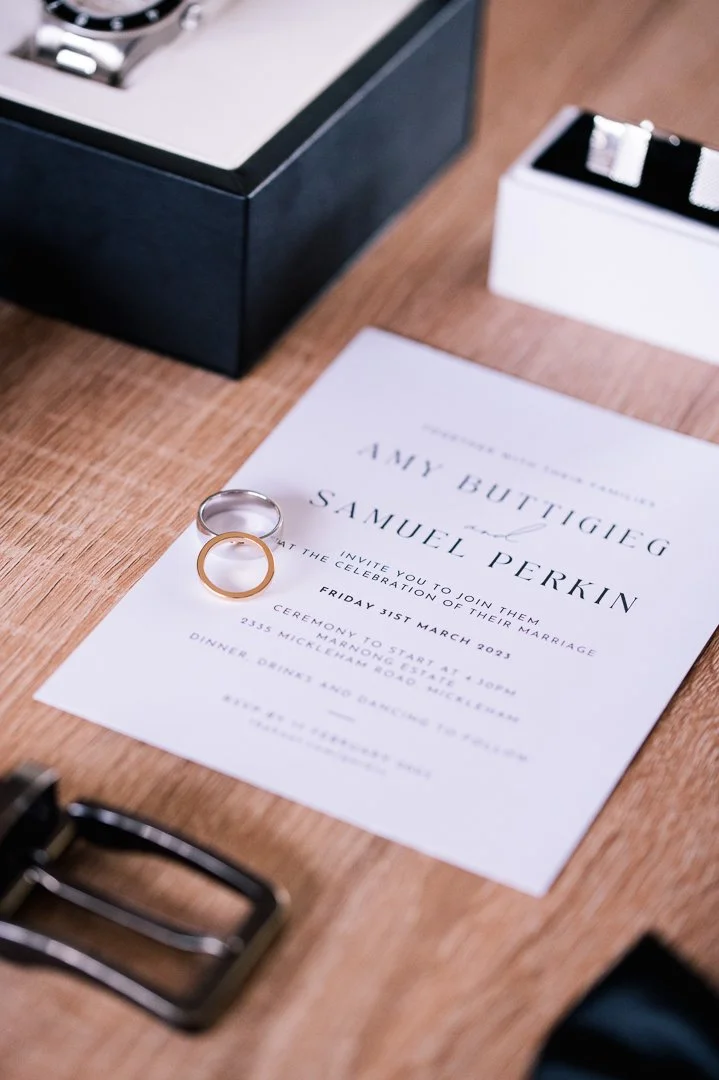 Wedding invitation for Amy Buttigieg and Samuel Perkin with rings on top, placed on a wooden surface near boxes and other accessories.