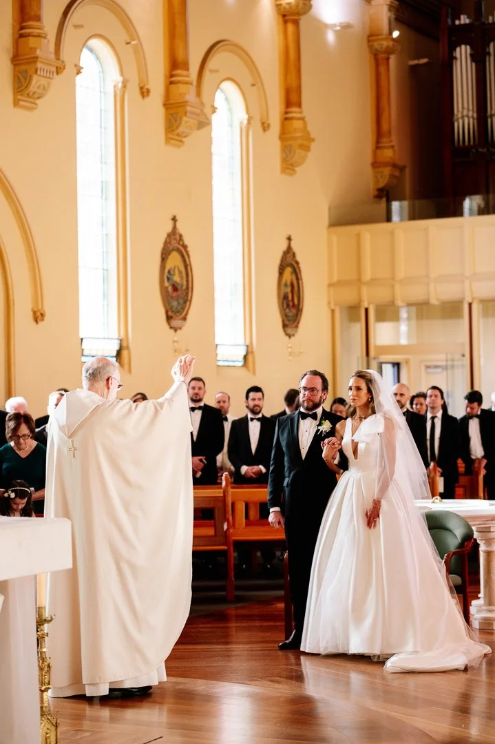A couple getting married at a church, holding hands and listening to the priest who is officiating the ceremony. The church has tall stained glass windows and ornate wood detailing.
