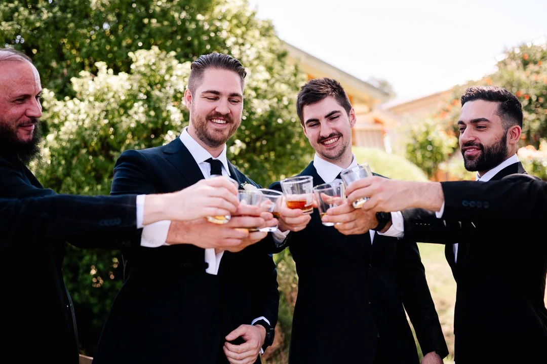 Four men in tuxedos, smiling and raising glasses of whiskey for a toast outdoors in a garden or yard.