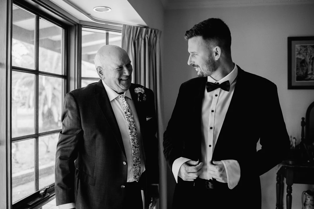 A young man in a tuxedo with a bowtie and an older man in a suit with a patterned tie share a joyful moment, smiling at each other indoors near a window.