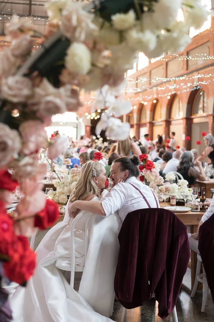 A bride and groom share a kiss at their wedding reception, surrounded by floral decorations and guests in a spacious, brick-arched venue with string lights.