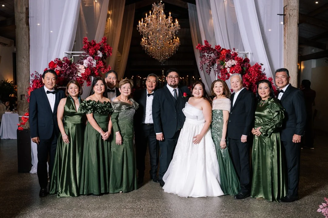 Group of people dressed in formal attire, standing in front of a floral wedding arch with a chandelier hanging above, at a wedding reception venue.