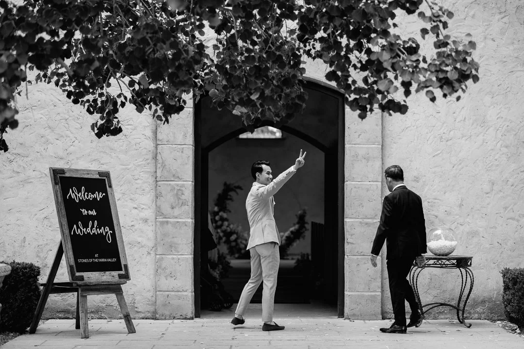 A black and white photo of two men in formal attire outside a wedding venue. One man is posing and making a peace sign, while the other is standing nearby. A signboard reads 'Welcome to our Wedding at Stones of the Yarrawar Valley' and a small table