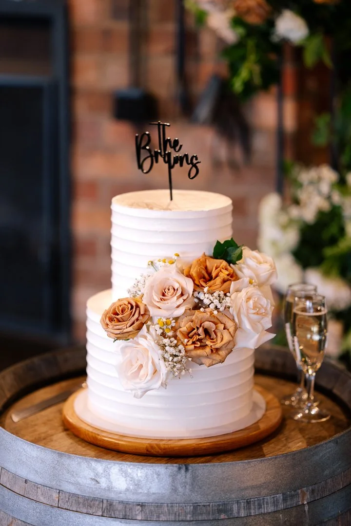 A two-tiered white wedding cake with floral decorations and a 'The Big Day' cake topper, placed on a wooden tray with a glass of champagne beside it.