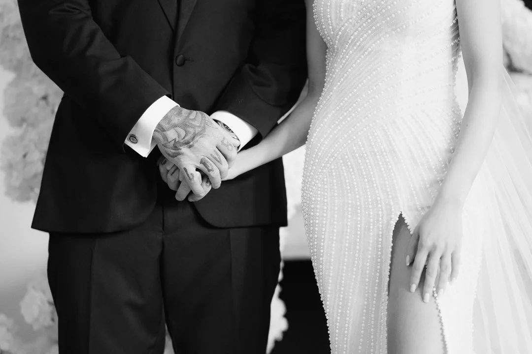 A bride and groom holding hands during their wedding ceremony, focusing on their joined hands and attire.