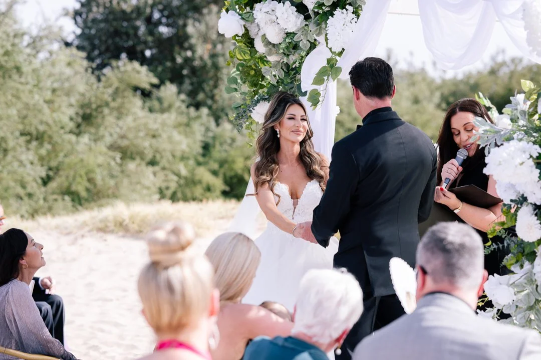 A bride and groom exchange vows at an outdoor beach wedding, with guests seated nearby under a floral arch.