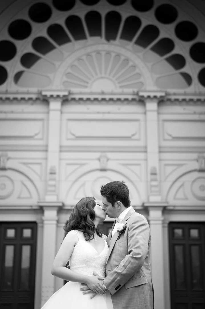 A black and white photograph of a bride and groom sharing a kiss, standing in front of a grand, ornate building with large windows and decorative architectural details.