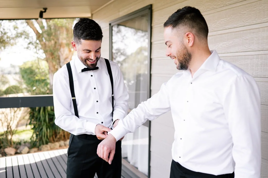 Two men in formal attire, one adjusting the other's sleeve outside a house with a wooden wall, greenery, and a deck in the background.