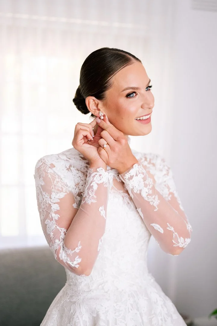 A bride with dark hair styled in an updo, wearing a lace wedding dress, adjusting her pearl earring.