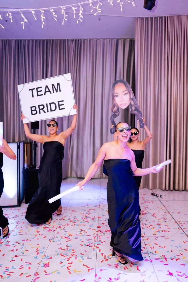 Women in black strapless dresses celebrating at a wedding reception. One woman holds a sign saying 'TEAM BRIDE', another holds a paper scroll, and a large photo of a woman's face is in the background. Confetti covers the floor.
