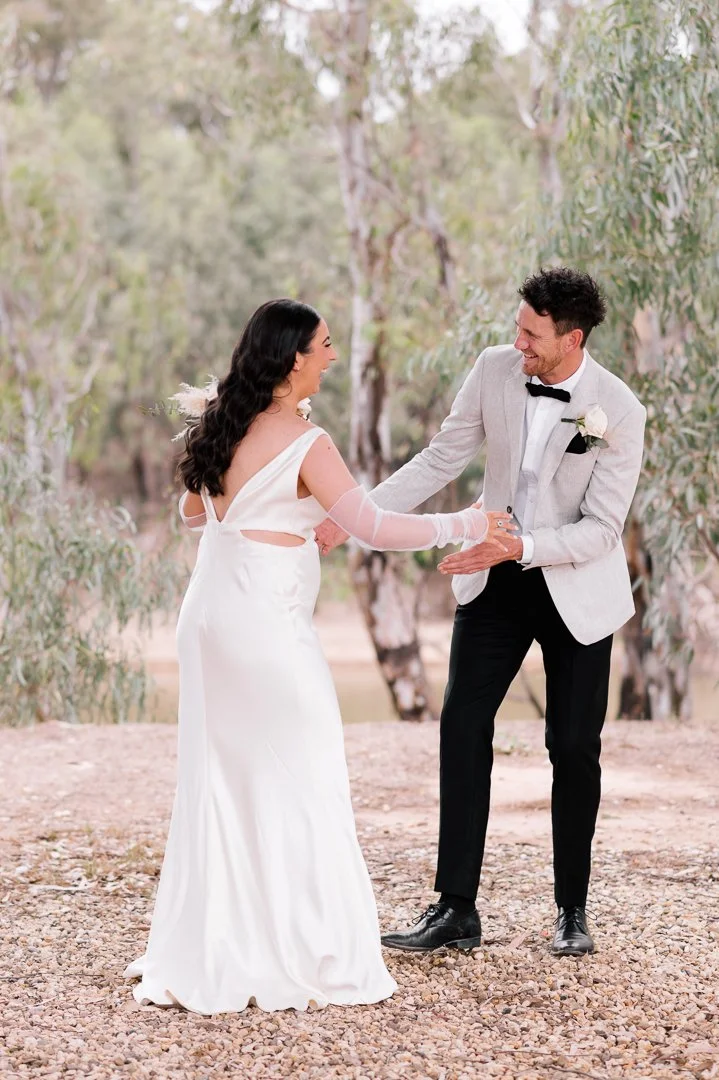 A bride and groom are smiling and holding hands outdoors in a wooded area, dressed in wedding attire, during their wedding ceremony or celebration.