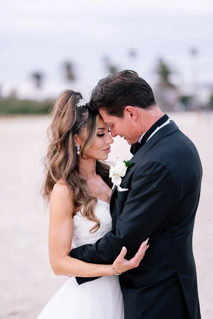 A bride and groom embracing on a beach, with their foreheads touching and eyes closed, during their wedding. The bride has long, wavy hair and is wearing a white wedding dress with jewelry, and the groom is in a black tuxedo with a white boutonniere.