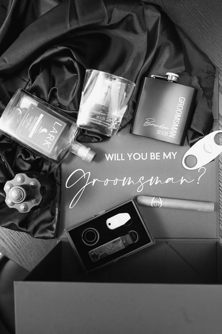 Various items including a glass, alcohol bottle, razor, gift box with a gift card, and other grooming products arranged on a dark fabric background.