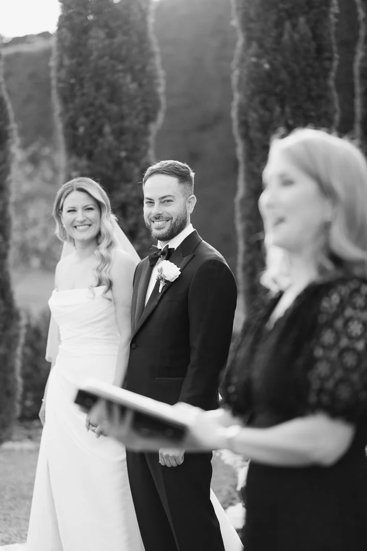 A black and white photo of a wedding ceremony outdoors. The bride and groom are smiling, facing the officiant who is holding a book, with tall trees in the background.