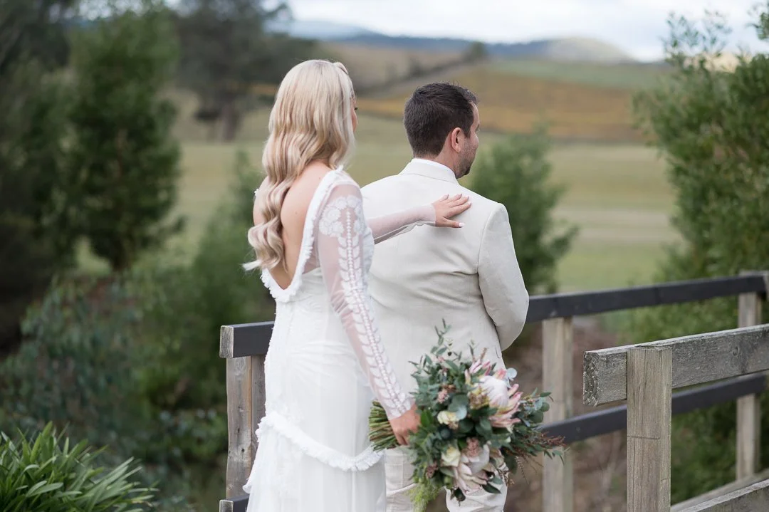 A bride and groom standing outdoors on a bridge, with the bride gently touching the groom's shoulder and holding a bouquet of flowers, overlooking a scenic countryside landscape.