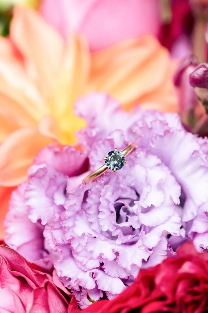 A close-up of a diamond solitaire ring resting on purple and pink ruffled flowers.