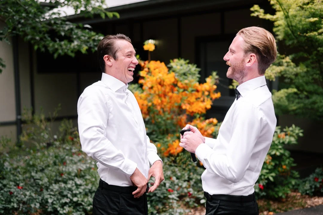 Two men in white dress shirts and black pants laughing and talking outdoors in front of a house with orange and green plants.