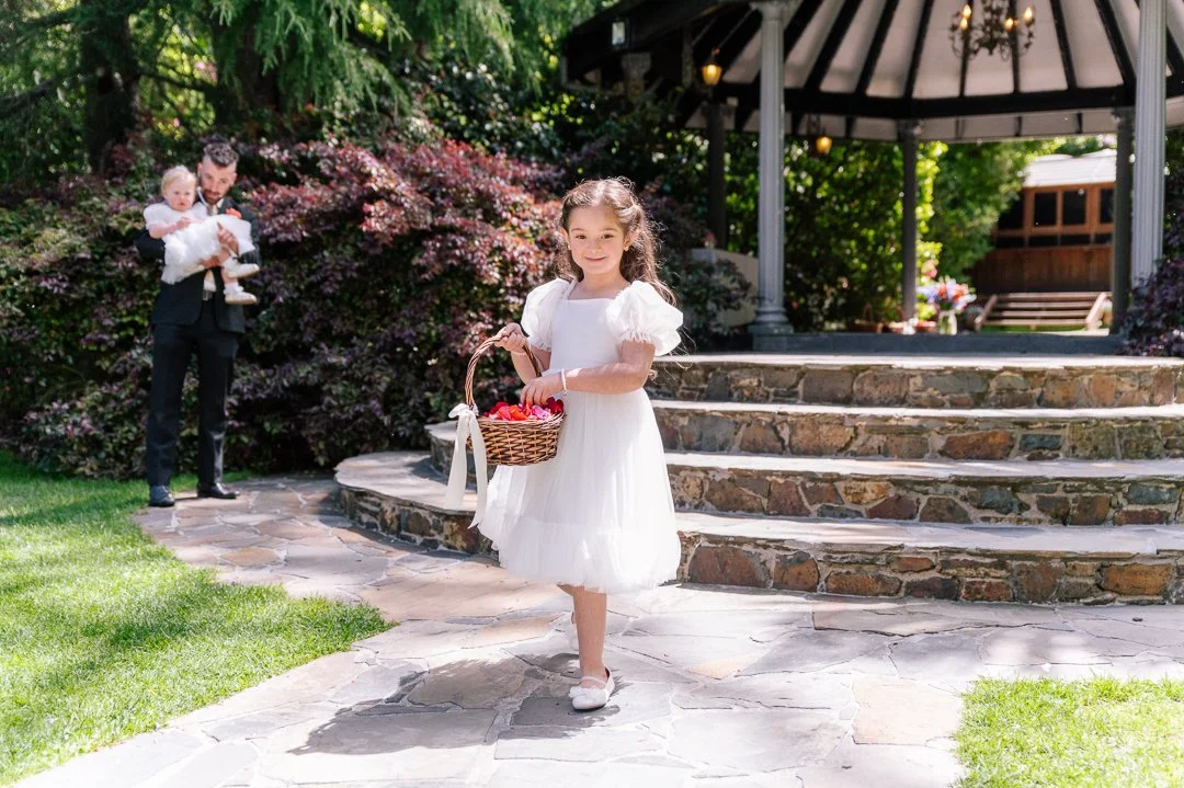 Young girl in a white dress holding a basket, standing outside on a stone patio near steps leading up to a gazebo, with a man holding a baby girl in the background.