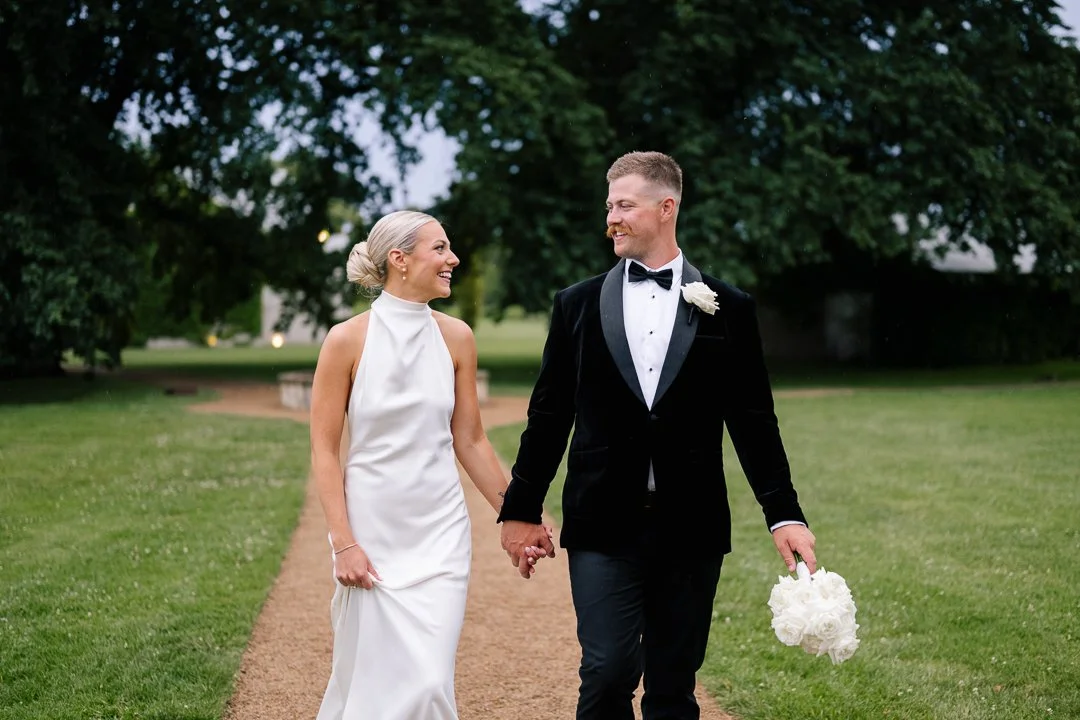Bride and groom walking hand in hand outdoors on a garden pathway, smiling at each other, with tall trees in the background.