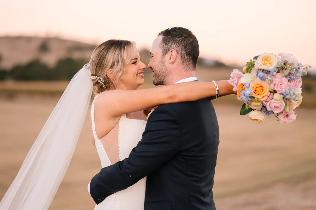 A bride and groom embrace outdoors at sunset, with the bride holding a colorful bouquet of flowers, smiling at each other.