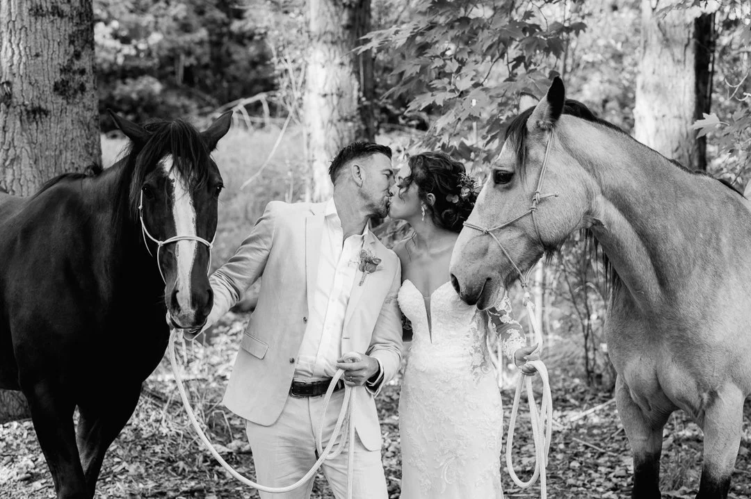 A black-and-white photo of a wedding couple kissing in a wooded outdoor setting, holding the reins of two horses on either side.