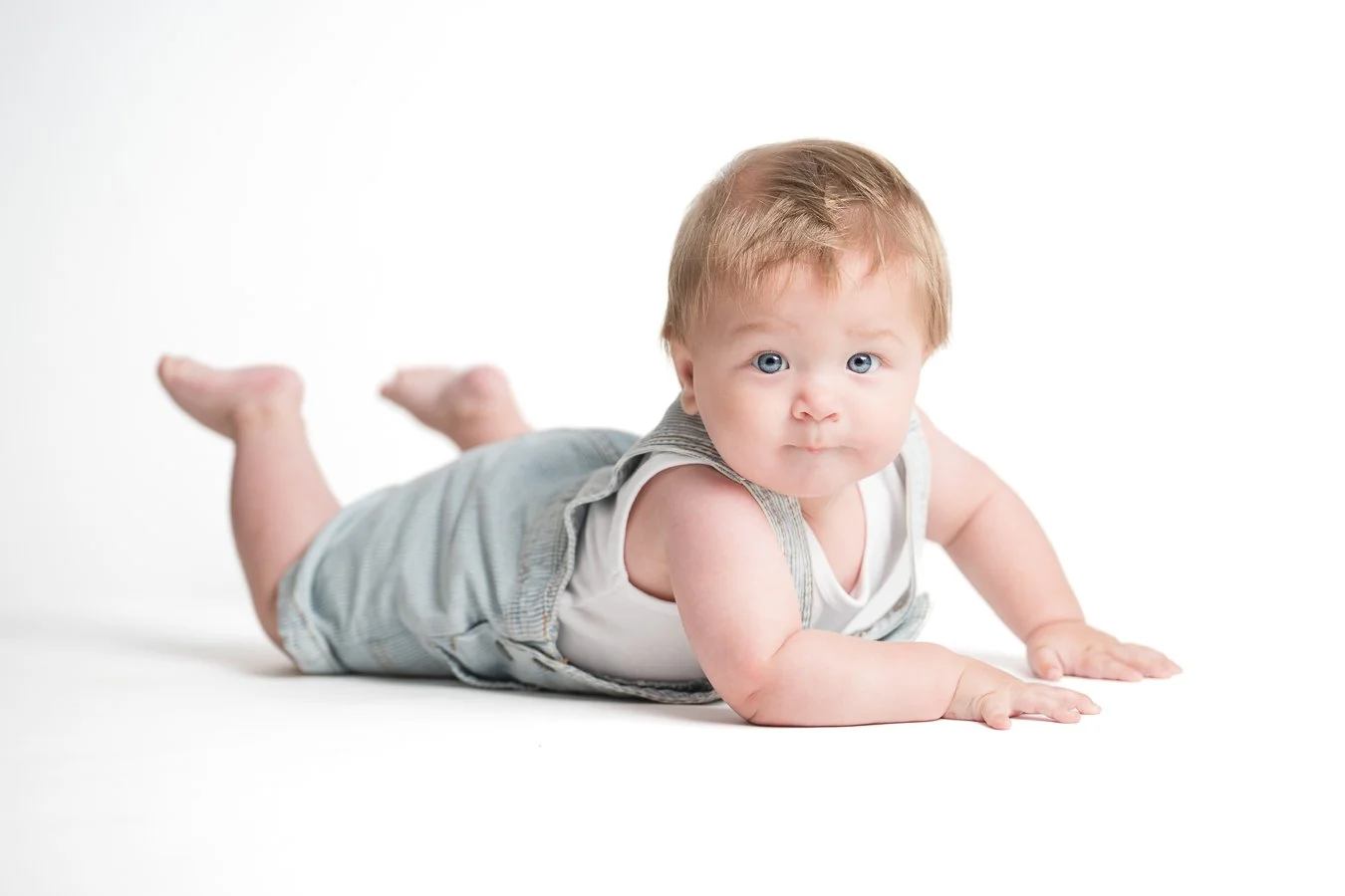 A baby crawling on a white surface, looking directly at the camera with blue eyes and light brown hair, wearing a gray sleeveless onesie.
