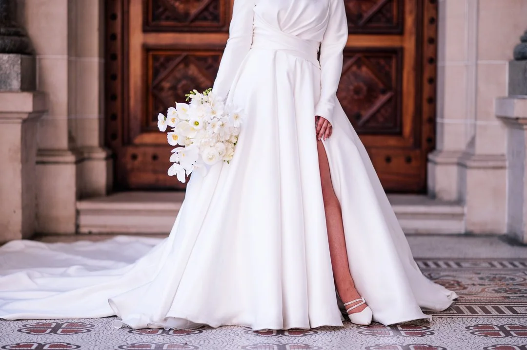 Bride in a white wedding dress holding a bouquet of white flowers, showing a slit revealing her leg, standing in front of a wooden door with stone surroundings.
