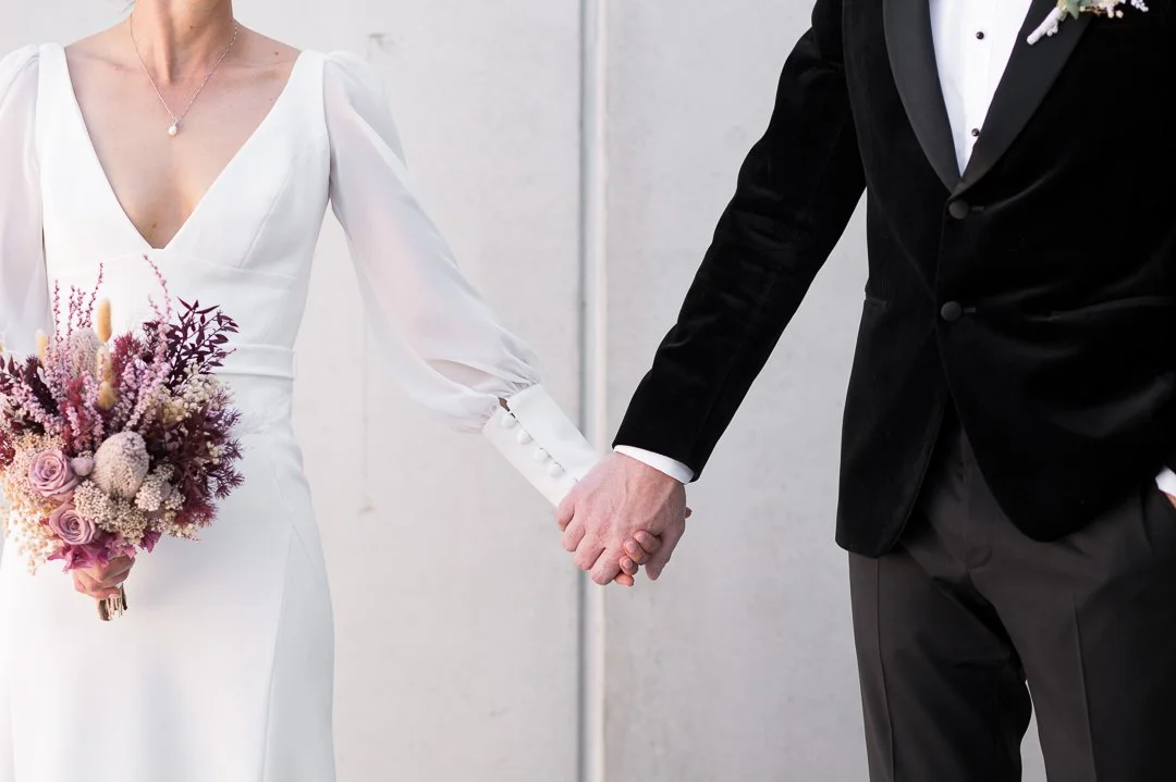 A bride and groom holding hands, dressed in wedding attire, with the bride in a white gown holding a bouquet of pink and purple flowers.