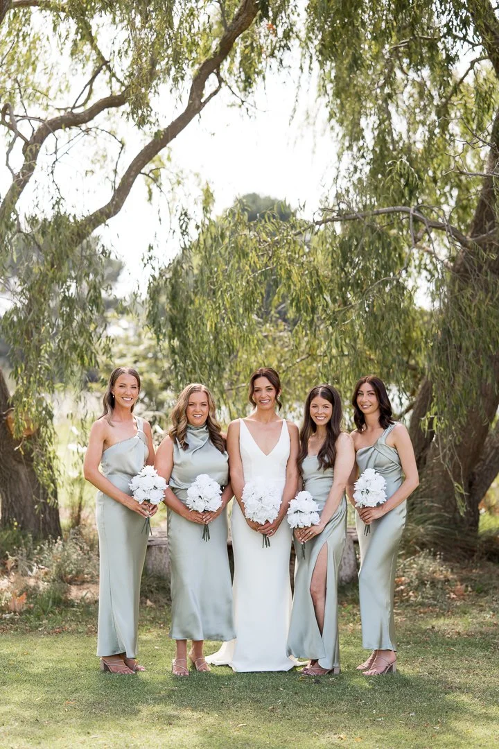 Bride and four bridesmaids standing outdoors under a large leafy tree, holding white bouquets, dressed in pastel-colored dresses.