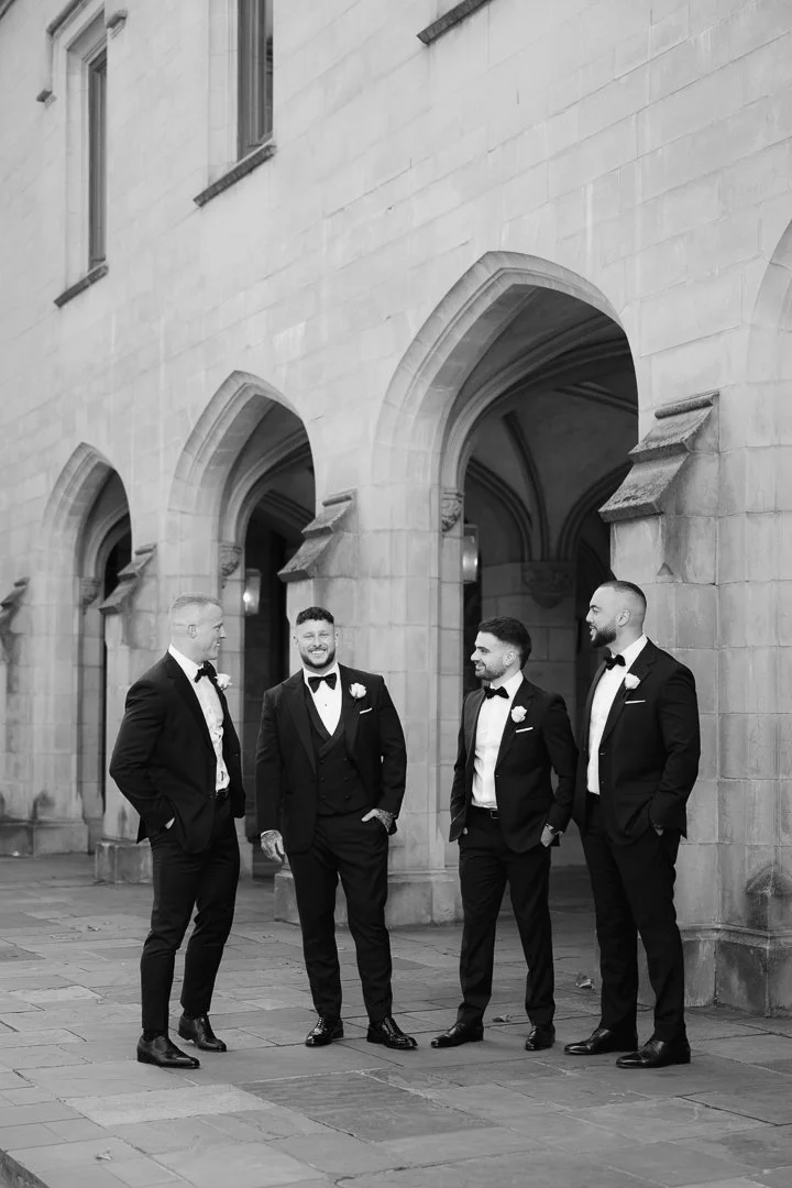 Four men dressed in tuxedos and bow ties standing and talking outside a building with arched doorways, in black and white.
