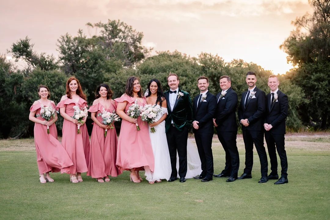 A wedding party standing outdoors on a grassy field at sunset, with nine people dressed in formal attire, including bride in white dress, groom in green velvet tuxedo, four bridesmaids in pink dresses, and four groomsmen in black suits.