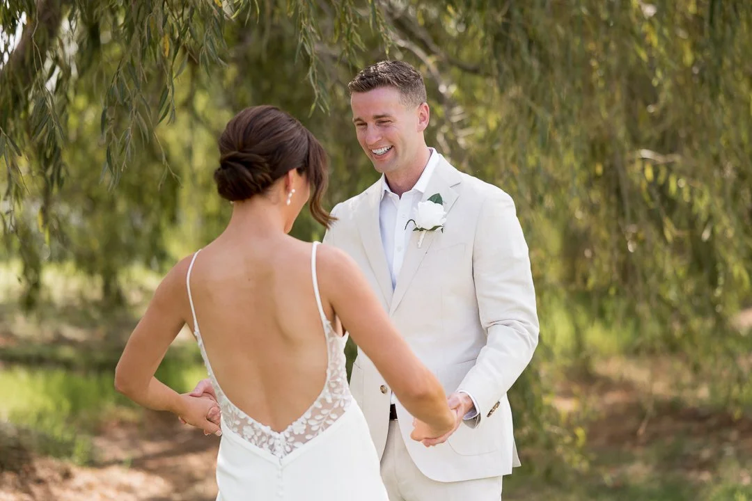 A bride and groom holding hands outdoors underneath trees, smiling at each other during their wedding.