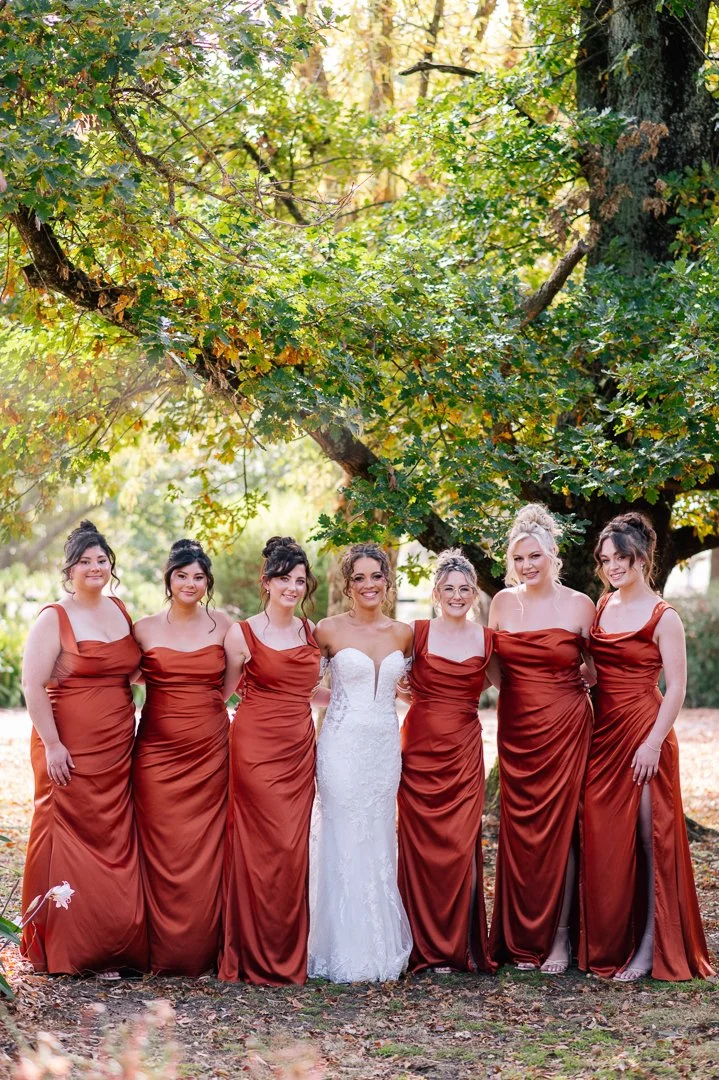 A bride in a white wedding dress standing with six bridesmaids in rust-colored gowns under a large leafy tree outdoors.