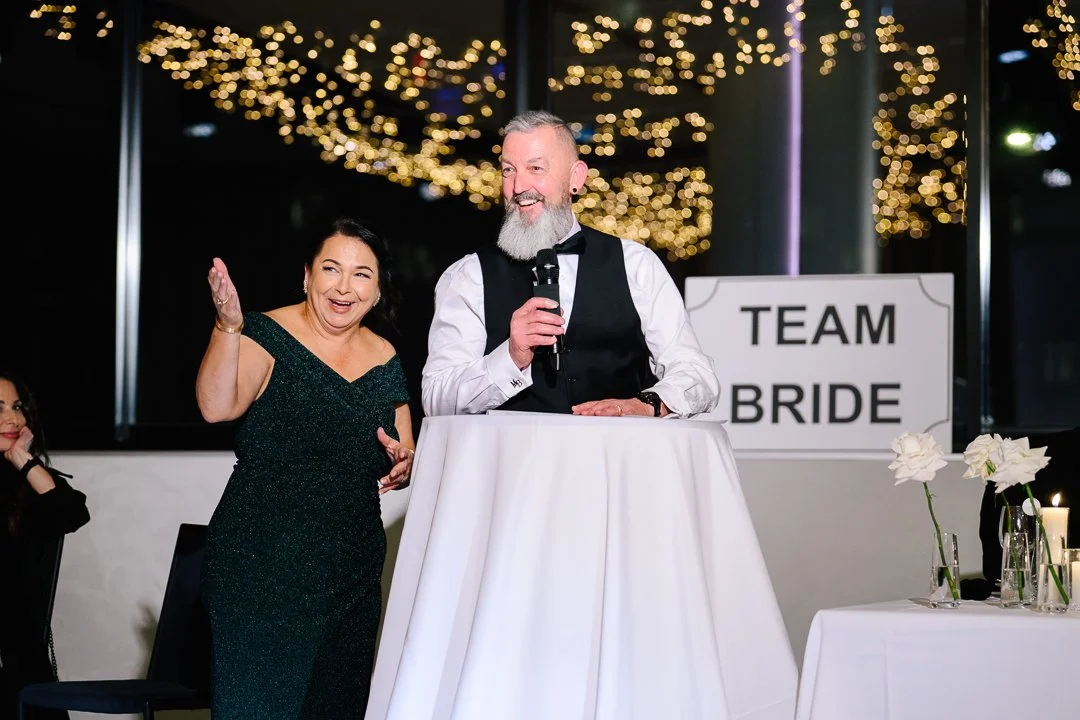 A man with a gray beard and a woman in a dark green dress smiling and speaking at a wedding reception. The man is holding a microphone, and a sign behind them reads 'Team Bride'. There are white flowers and candles on a table nearby, and decorative s