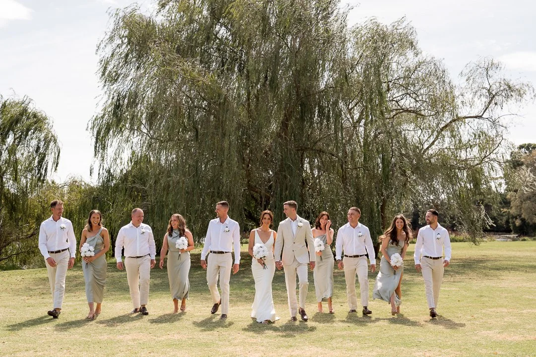 A wedding party walking on grass in front of large trees, with the bride and groom in the center and eight bridesmaids and groomsmen on either side.